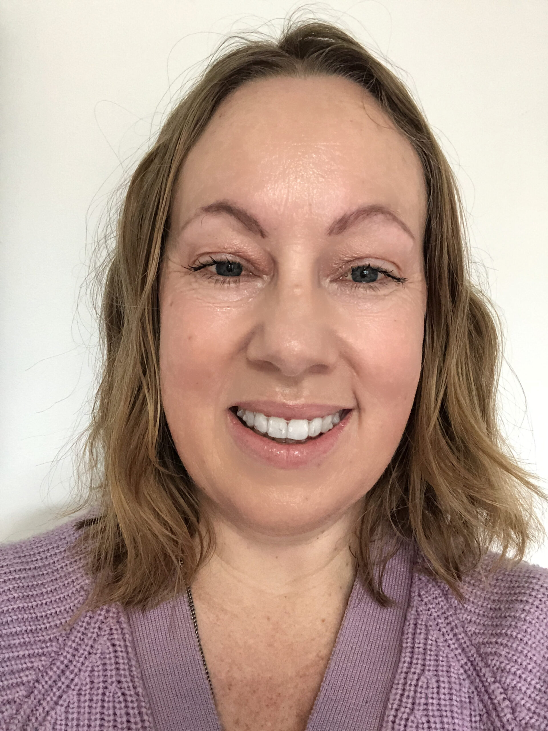 A woman with a dark blonde wavy lob haircut, with a smile and wearing a lavender knit top